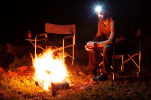 night time photo of man with headlamp on, creating light burst, sitting by fire on camping chair with another chair next to him