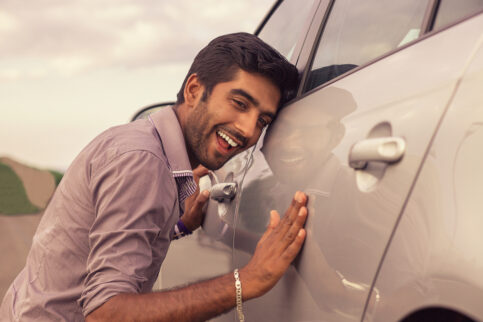 Young hispanic man wearing in formal shirt holding is petting his car and smiling outdoors. A new car, new driver concept