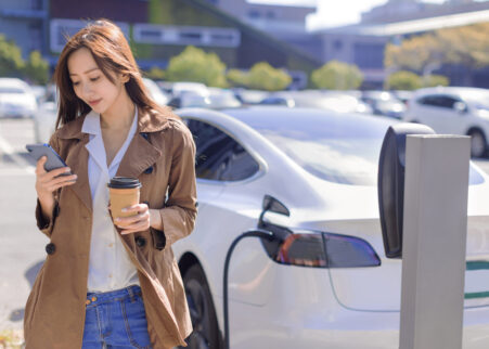 Smiling young woman standing on city parking near electric car, charging automobile battery from small city station, drinking coffee and using smartphone